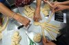 Vue du dessus d'une table couverte de pâtons de pâte à pain en train de prendre forme. Trois paires de mains s'affairent pour façonner ces pains en forme de femmes bretzels
