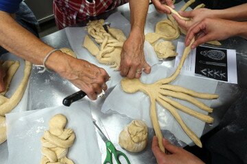 Vue du dessus d'une table couverte de pâtons de pâte à pain en train de prendre forme. Trois paires de mains s'affairent pour façonner ces pains en forme de femmes bretzels